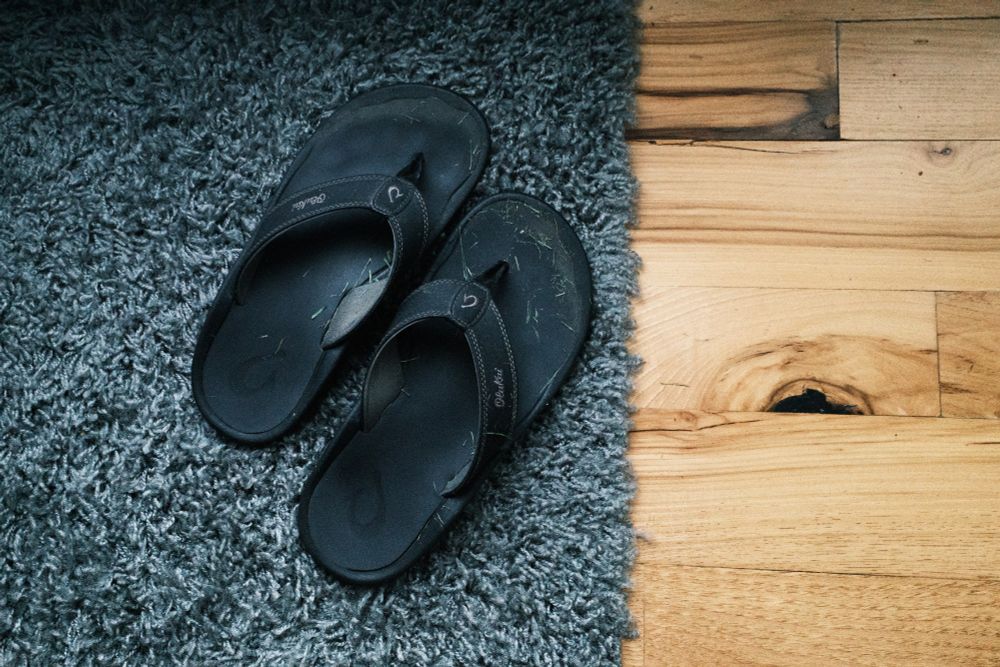 A pair of Olukai flip flop shoes sit on a small dark teal colored rug with a wooden floor on the right third of the image. The flip flops have green grass clippings on the black sole of the shoe. The small rug occupies 2/3rds of the image.