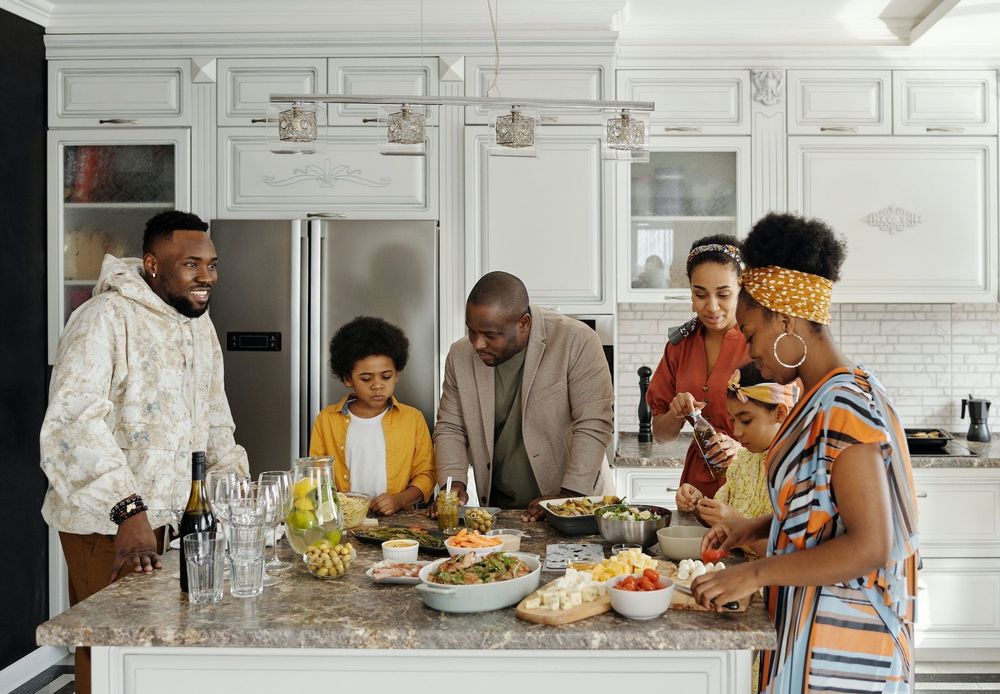 A family of six gathered around an island in a kitchen with food on it.