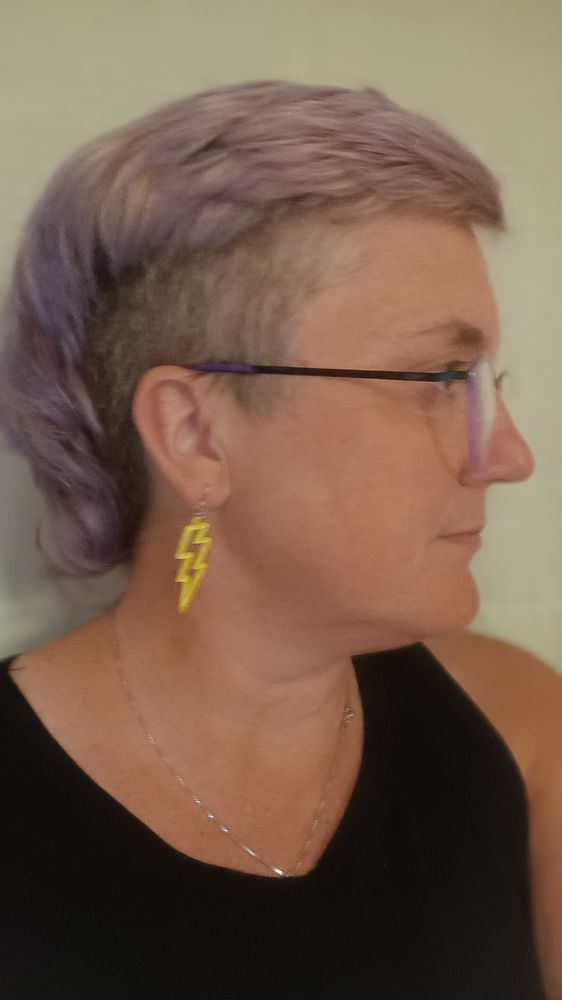 Selfie mit kurzen lila Haaren und Sidecut, lila Brille und gelbe Blitzohrringe, schwarzes Unterhemd, ich schaue zur Seite, ernst