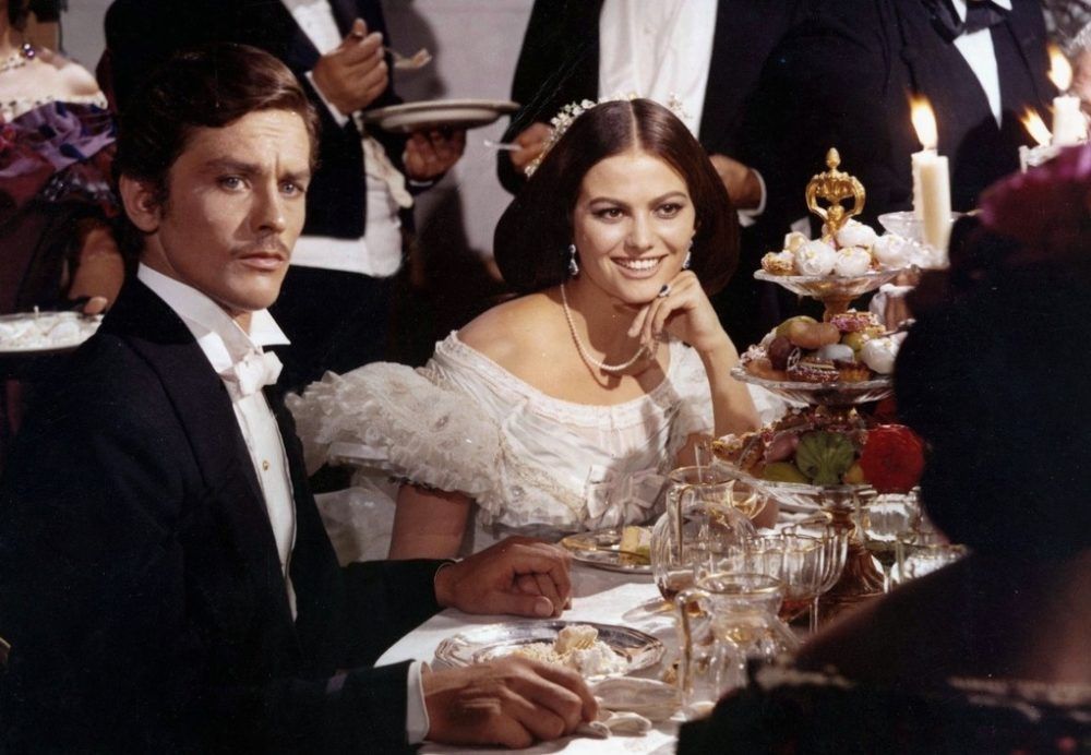 Screen grab from The Leopard showing a man and woman in fancy dress and seated at a table. The actress playing the woman is Claudia Cardinale.