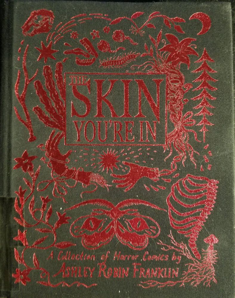 cover to The Skin You're In by Ashley Robin Franklin
the cover is black cloth with red metallic ink showing the title and assorted body parts, bones, bugs and plants arranged in a pleasing manner