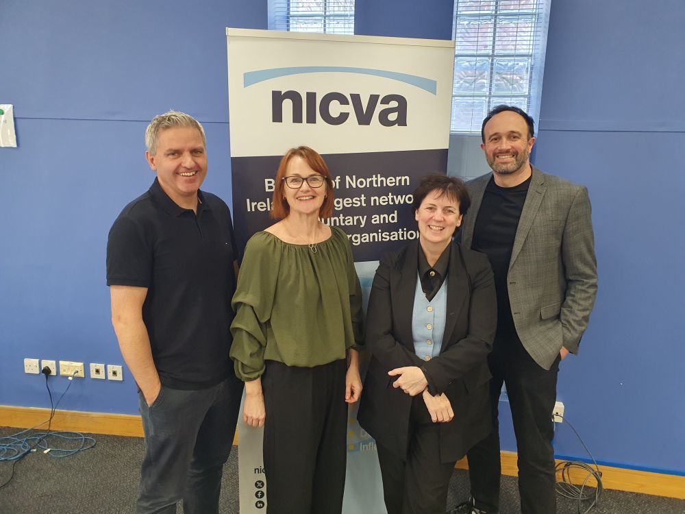 Photo of Adrian Johnson, Innovation Commissioner, Sandra Bailie, NICVA's Head of Organisational Development, Roisin Kelly, NICVA's Skills Development Coordinator, and Stephen Dallas, CEO of The Bytes Project. They are standing together, smiling to the camera, in front of a NICVA popup banner.