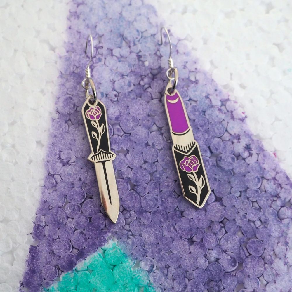 A shop photo from Wild Fancy Designs of their dagger + lipstick earrings. The earrings are a golden brassy color. One is a dagger with a rose design on the handle in mauve. The other is a lipstick in the same mauve, with the same rose design on the case.