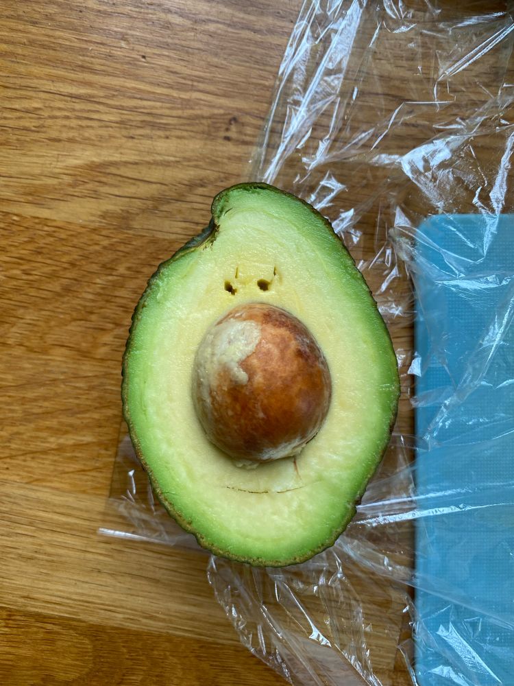 Half an avocado, the stone in the centre looks like a large nose while there are blemishes in the flesh that look like two small eyes and a slightly smiling mouth. A dreamworld-esque avocado. 
