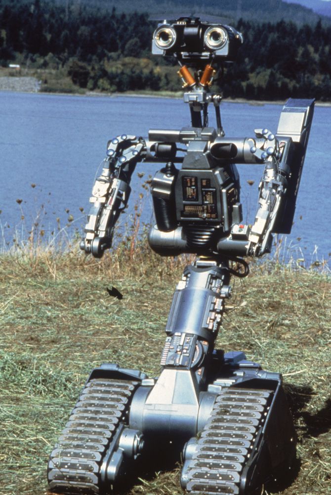The robot, Johnny Five, from the 1988 film ‘Short Circuit’. He looks like Wall-e’s taller, skinnier dad. A short, wide boxy head, with large camera lens eyes, a vaguely humanoid torso (complete with shoulder mounted laser) and a narrow body. He moves around on two caterpillar tracks. In this photo he’s standing outside on grass, with a body of water behind him. 