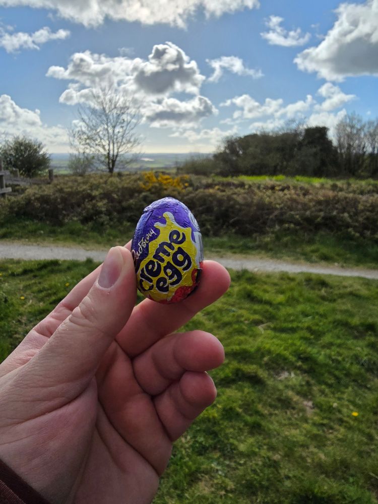 Hand holding an unopened Cadbury Creme Egg on a trail in the hills of the Cornish countryside in England.