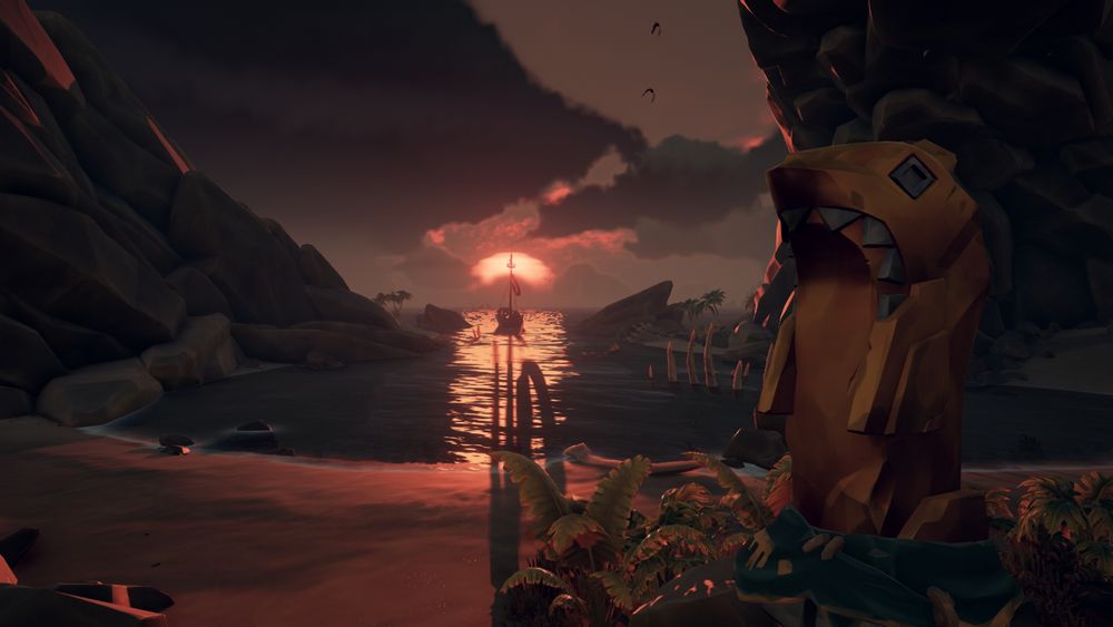 A photo taken from the perspective of standing on a beach looking across to steep rocks on either side of an open cove. Ahead in the distance floats a boat silhouetted by a sunset on the horizon as clouds gather above. To the right side, close to the camera and perched atop a rocky shrubbed area stands a durpy looking gold shark statuette 