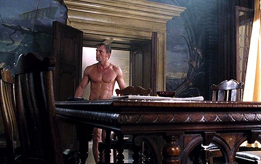 A nude Daniel Craig walks up to a large ornate wooden table covering his crotch in a fancy Italian apartment and lift a pistol and ammo while looking around 