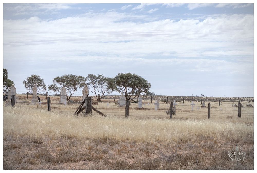 A colour photograph of a remote, sparsely populated outback cemetery on a clear but cloudy day. The foreground is dominated by dry, pale yellow and tan wild grass and arid, reddish-brown dirt.

A dilapidated, low wire fence with thick wooden posts runs across the middle ground. Beyond the fence, several pale headstones and monuments are visible, interspersed with a few small, round-canopied green trees.

The land appears flat and extends to the horizon under a vast, pale blue and white sky filled with light cirrus clouds.