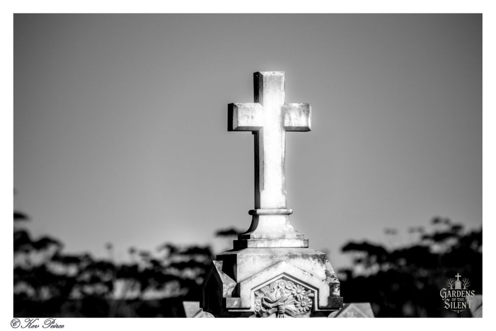 A black and white, low angle photograph focusing on a weathered, stone Celtic cross monument standing atop a decorated plinth in Kapunda St. Johns cemetery.

The cross is brightly lit, contrasting sharply with the nearly white sky behind it. The base of the monument features a carved floral element.

The background is softly blurred, showing the dark, silhouetted shapes of distant trees and bushes beneath the bright sky.