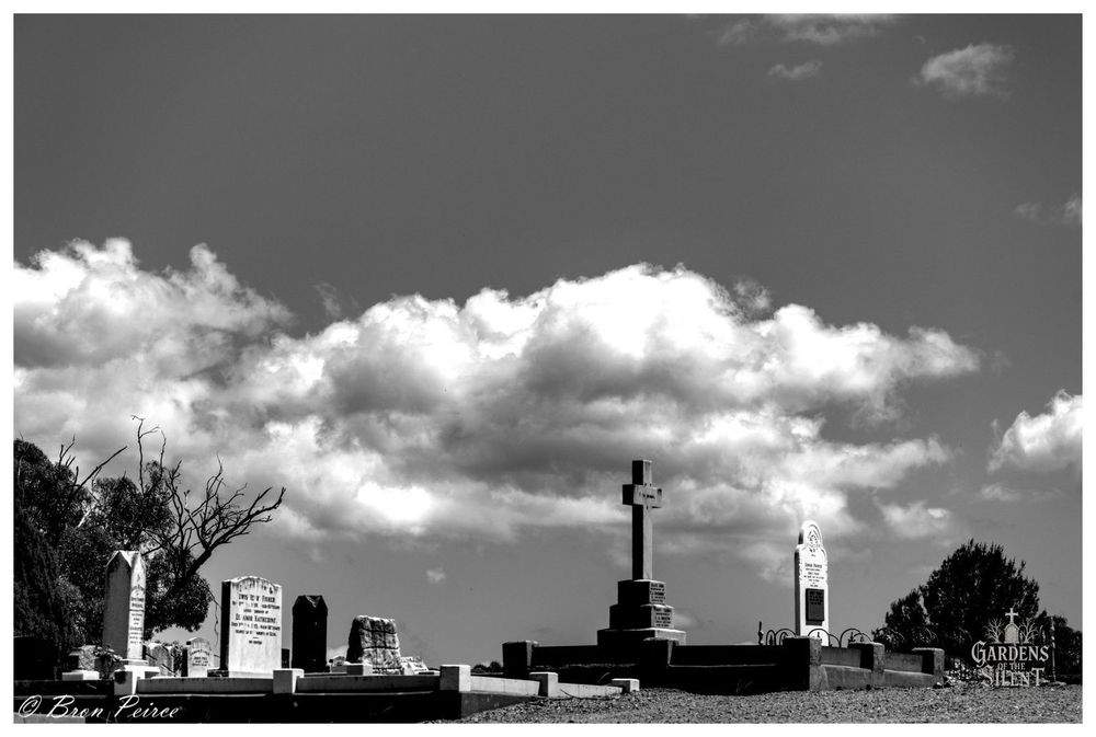 Black and white low angle photograph of several grave markers in the Wirrabara Cemetery, signed by Bron Peirce. The central feature is a large, square stone cross on a tiered base, positioned slightly right of centre.

To the left are several other weathered headstones, one of which is a tall, white marble tablet. The scene is dominated by a dramatic, bright sky filled with large, billowing cumulus clouds that stretch across the horizontal center, appearing luminous against the dark sky

A few bare tree branches are visible on the far left, and dark foliage frames the scene on the right.
