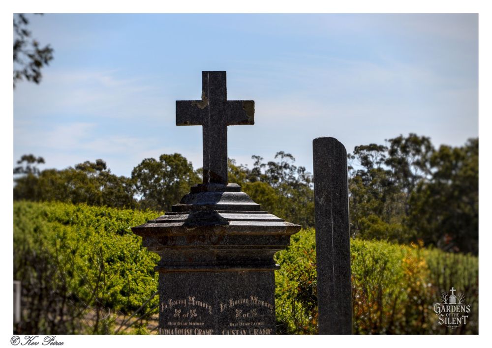 A dark stone cross monument set on a thick pedestal, silhouetted against a bright blue sky.

The base has visible inscriptions. Behind the monument is a vibrant green vineyard and dense line of eucalyptus trees.