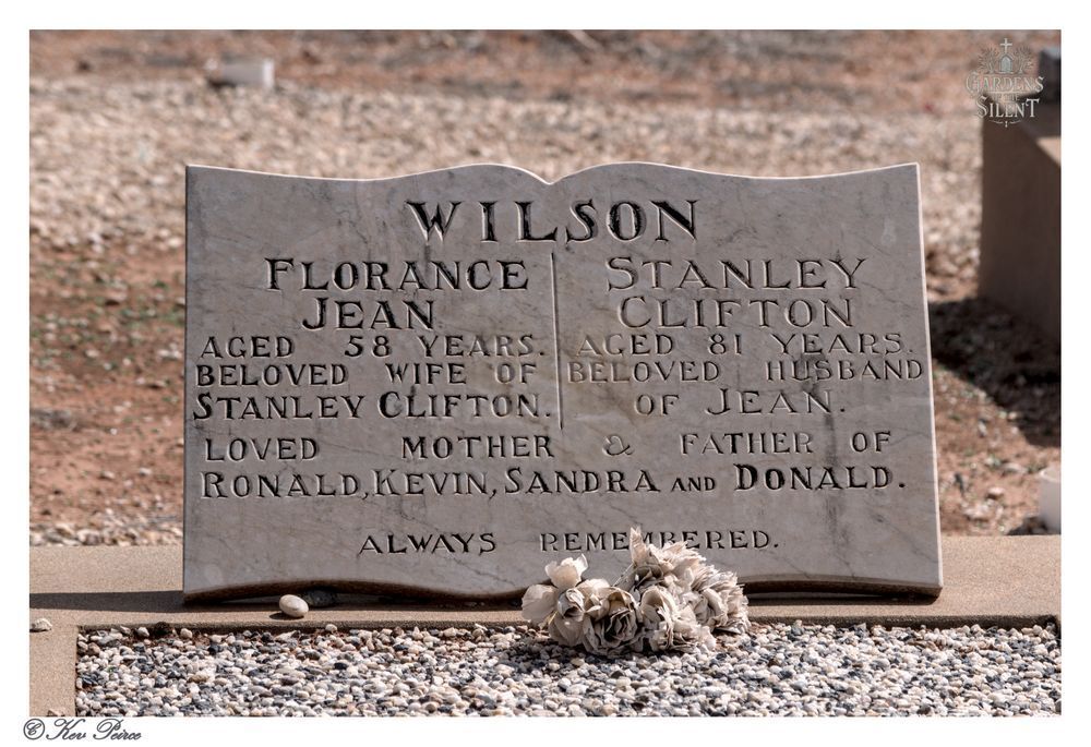 A modern, light coloured marble headstone for the Wilson family, located at Berri Cemetery.

The stone is carved in a scroll-top style and is dedicated to FLORANCE JEAN (Aged 58 years, Beloved Wife of Stanley Clifton) and STANLEY CLIFTON (Aged 81 years, Beloved Husband of Jean).

The bottom inscription reads: "LOVED MOTHER & FATHER OF RONALD, KEVIN, SANDRA and DONALD. ALWAYS REMEMBERED."

A small cluster of dried white or pale flowers rests on the light-colored gravel in front of the stone.