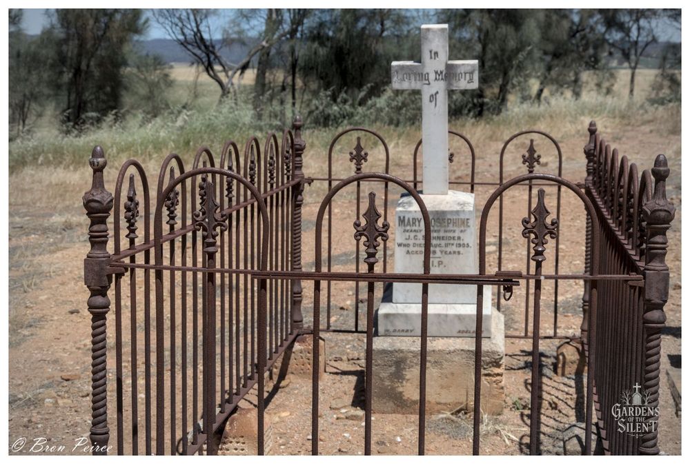 A photograph signed by Bron Peirce. The central focus is a historic grave surrounded by a rusty, ornate cast iron fence.

Inside the enclosure, a white marble headstone is mounted on a rough concrete plinth. The headstone is topped with a simple cross.

The grave is located in dry, flat ground typical of the Australian bush, with sparse green and dead grasses and some distant scrub and trees under a hazy white sky.