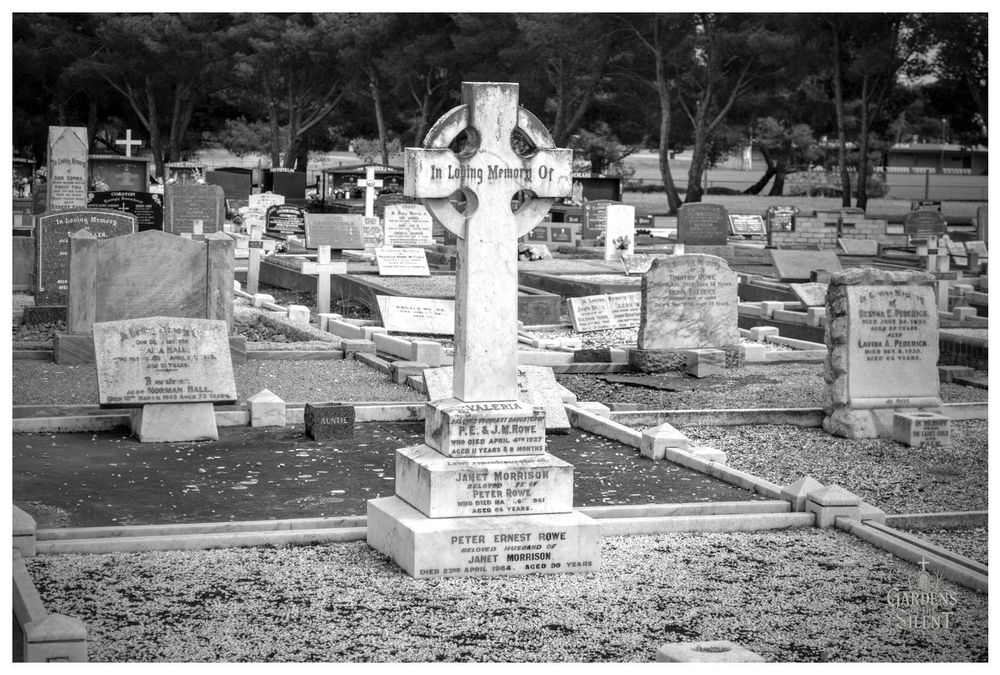 A central black and white photograph of a cemetery plot featuring a prominent white stone monument in the shape of a traditional Celtic cross. The cross stands on a multi-tiered square base.

The plot is surrounded by low marble curbing and filled with light coloured gravel. Numerous other gravestones are visible in the background, which is lined with dark trees.