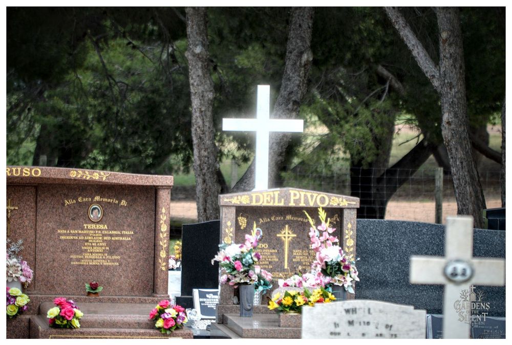 A mid distance photograph of several modern, polished granite monuments in the Two Wells Cemetery, framed by dark pine trees and foliage.

The central memorial, a dark brown headstone features a white cross at its top and two prominent floral wreaths one on the left and one on the right, decorated with pink, yellow, and white flowers.

To the left is a large, brown granite monument with a small framed photo. The overall scene has a softer focus, suggesting a quiet, established, and well maintained area of the cemetery.