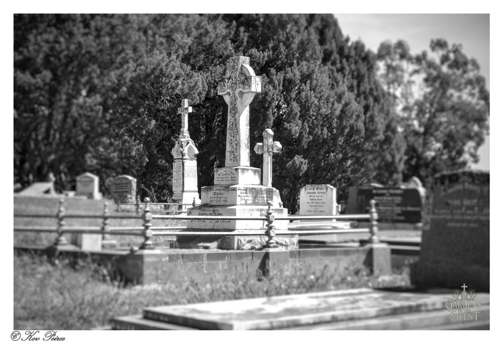 A striking black and white, focus stacked photograph capturing a central, highly ornate Celtic cross headstone monument in the Rowland Flat Cemetery.

The large cross, which has detailed knotwork, is flanked by two smaller stone crosses, and all three are dramatically framed against a dense, dark backdrop of trees.

The main monument rests on a dark, tiered foundation and is partially surrounded by a low fence or railing. The foreground, which is intentionally blurred, consists of grass and the tops of other grave markers, drawing the viewer's eye to the towering central memorial.

The high contrast enhances the solemn, historic mood of the scene. Signed Kev Peirce.