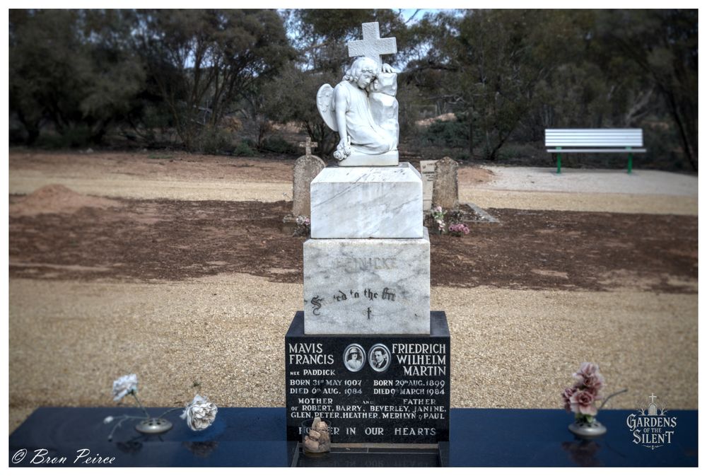 A tall marble headstone topped with a white marble statue of a seated angel leaning its head against a cross. The base of the monument is made of dark stone with inscriptions and two small framed portrait photos.

The inscription names are MAVIS FRANCIS PADDICK and FRIEDRICH WILHELM MARTIN. The ground around the grave is bare earth and wood chips, leading to a natural, treed background.

A white park bench sits to the right in the distance. Small, faded flowers are placed near the base. Photo by Bron Peirce.