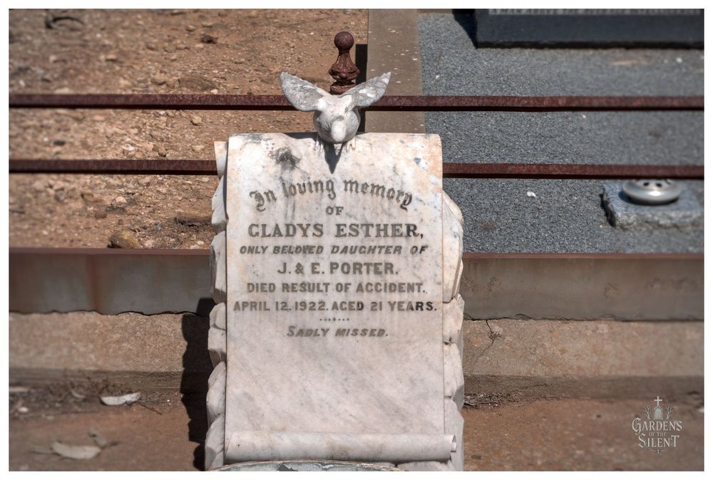Close up, focused photograph of a small, vertical marble gravestone in the Wirrabara Cemetery. The marker is shaped like a scroll and is topped with a weathered, possibly dove or angel wing-shaped stone carving and a small rusted finial.

The inscription reads: 'In loving memory of GLADYS ESTHER, ONLY BELOVED DAUGHTER OF J. & E. PORTER. DIED RESULT OF ACCIDENT. APRIL 12, 1922. AGED 21 YEARS. SADLY MISSED.'

The stone is set into a concrete border with dry, reddish brown earth and two horizontal rusty metal bars visible behind it.