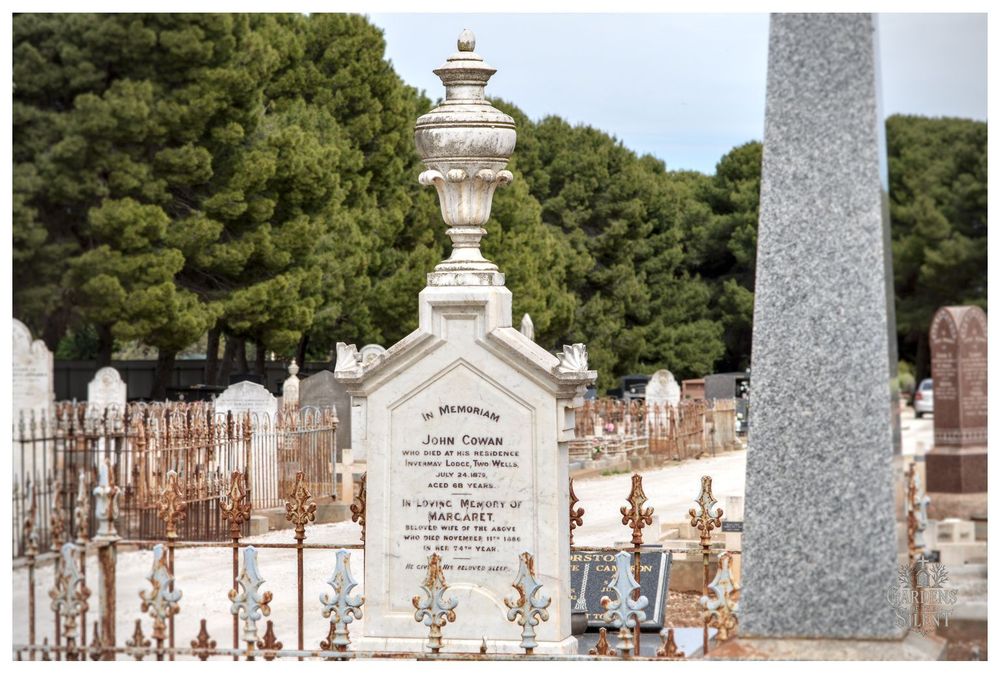 An ornate, white marble gravestone in a cemetery, featuring a large, decorative funerary urn on top of a base shaped like a house gable.

The monument is surrounded by a low, rusted, decorative iron fence. Other older gravestones and a tall, grey granite obelisk are visible in the foreground. A line of pine trees runs across the background.
