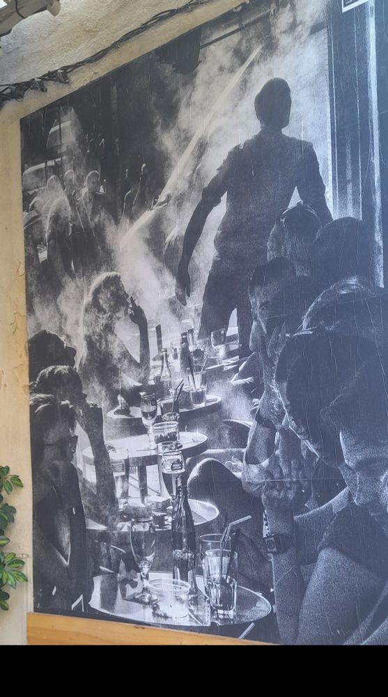 Photo d'un mur de rue où se trouve une grande photo noir et blanc d'un groupe de personnes qui qui boivent un coup au bar.
Auteur non mentionné