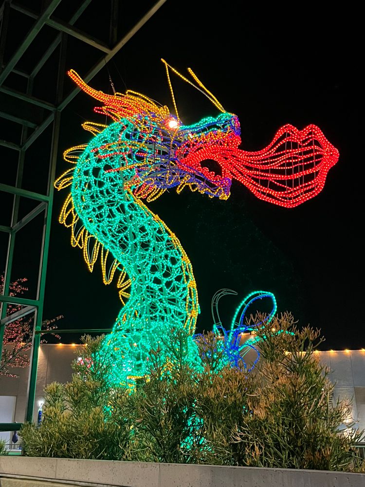 A green dragon created from string lights, the photo shows only its neck and head, rearing back and blowing red flame 