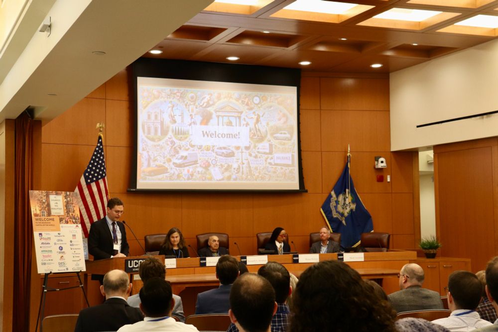 A man stands at a podium addressing an audience in a conference room, with a "Welcome!" slide displayed on a screen behind a panel of four seated speakers.