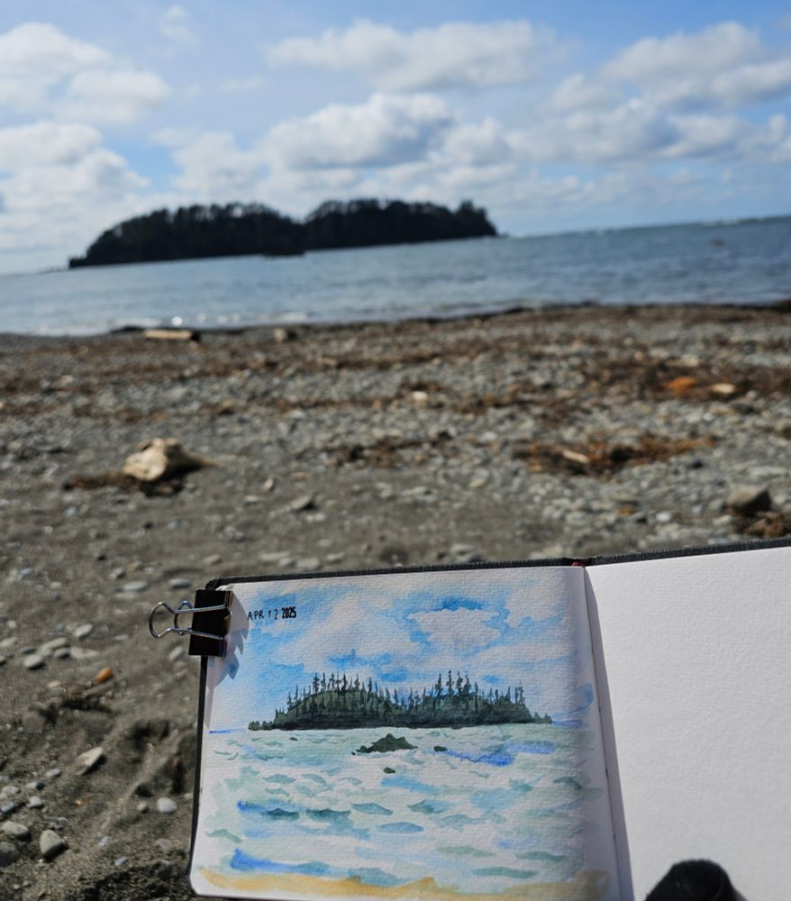 Photograph of a sketchbook being held against a coastal backdrop. The weather is partly cloudy, and a large island can be seen in the distance. The sketchbook shows a watercolor sketch of the ocean and island. 