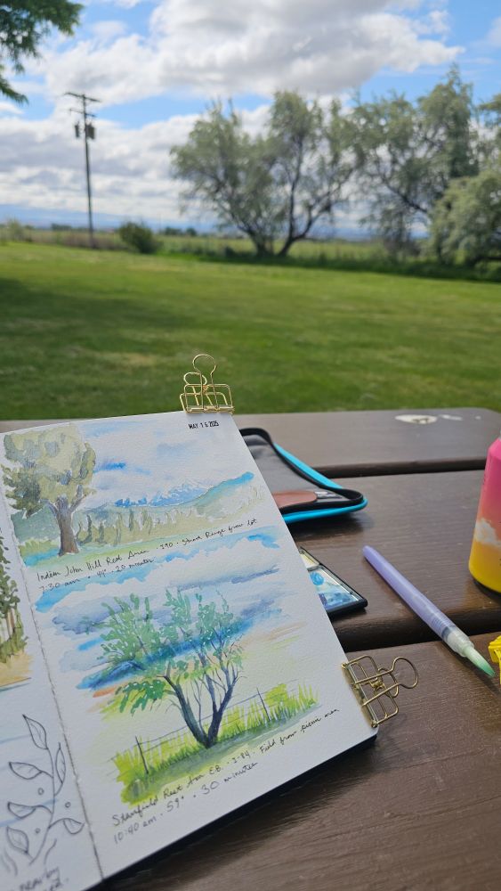 Sketchbook held up on a picnic table. The page has two sketches of two different rest area scenes with trees, both done in 20-30 minutes. 