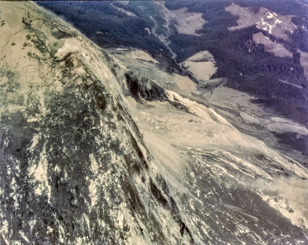 Aerial view of the initial north flank landslide on Mount St. Helens. Slide blocks from the bulge drop away and a gray dust sheet rides downslope toward the forested valley and clearcuts. Dorothy Stoffel. May 18, 1980. About 8:32–8:33 a.m.