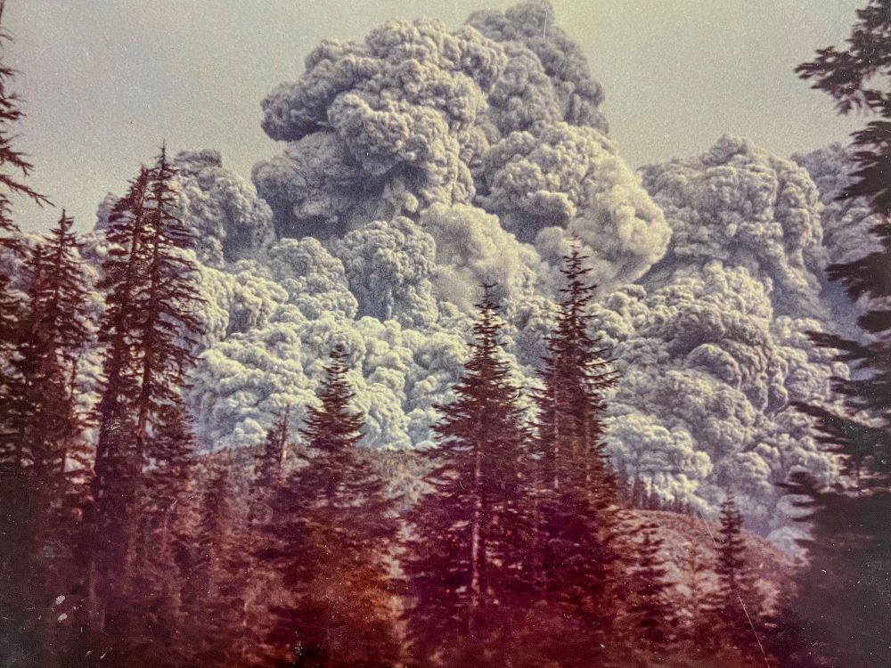 The eruption intensifies as a dense ash cloud roils into the sky, stretching above the tree line. Steam and ash pour from the breached summit, with mushrooming plumes forming behind the southern ridges. The flanks of the mountain are covered by ash.

Photo by Kevin Millican.