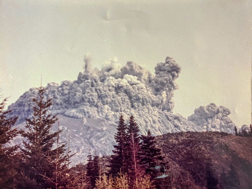 An ash cloud begins to rise from Mount St. Helens shortly after 8:32 a.m., as seen from the south side. The eruption column billows upward as dark material shoots skyward, framed by evergreens in the foreground. A lower plume to the right is likely connected to the lateral eruption.

Photo by Kevin Millican.