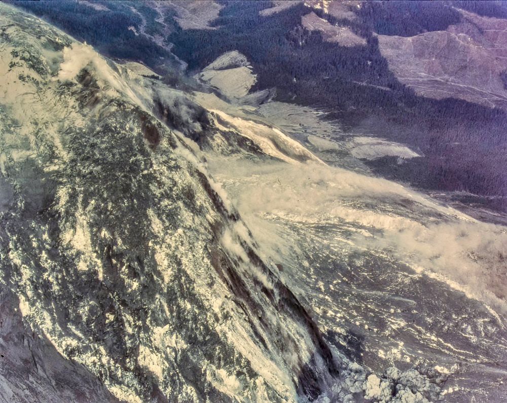 Wider view of the debris avalanche accelerating off the north side. Elongate slide blocks and fresh scarps stripe the slope as a ground-hugging dust cloud marks the moving mass into the North Fork Toutle valley. A small plume of ash and steam appears at the bottom-right margin. Dorothy Stoffel. May 18, 1980. About 8:33 a.m.