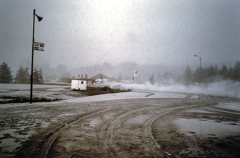ChatGPT said:

View of a Chevron gas station in Morton, Washington, around 12:28 p.m. on May 18, 1980, blanketed in ash from the Mount St. Helens eruption. Through the haze, a Puget Sound Bank building is visible in the distance, while tire tracks curve across the ash-covered pavement in the foreground. A passing vehicle has thrown up a plume of fine gray dust, forming a “rooster tail” across the road.
Photo by Keith Ronnholm. Uploaded by Steven Rosenow on 1980 Eruption of Mount St. Helens Facebook group.
