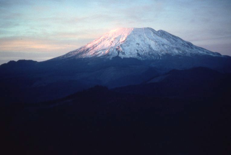 Mount St. Helens at sunset on Oct. 27, 1980, its upper slopes covered in snow and glowing faintly pink in the evening light. The lower ridges are in shadow under a calm, partly cloudy sky. Skamania County, Washington. USGS photo.