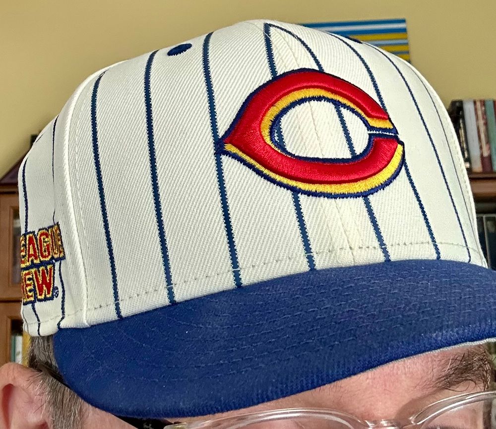 Cincinnati Reds baseball hat with blue visor, white dome with blue pinstripes. red C logo with yellow drop shadow all outlined in black. Big League Chew logo on one side. 