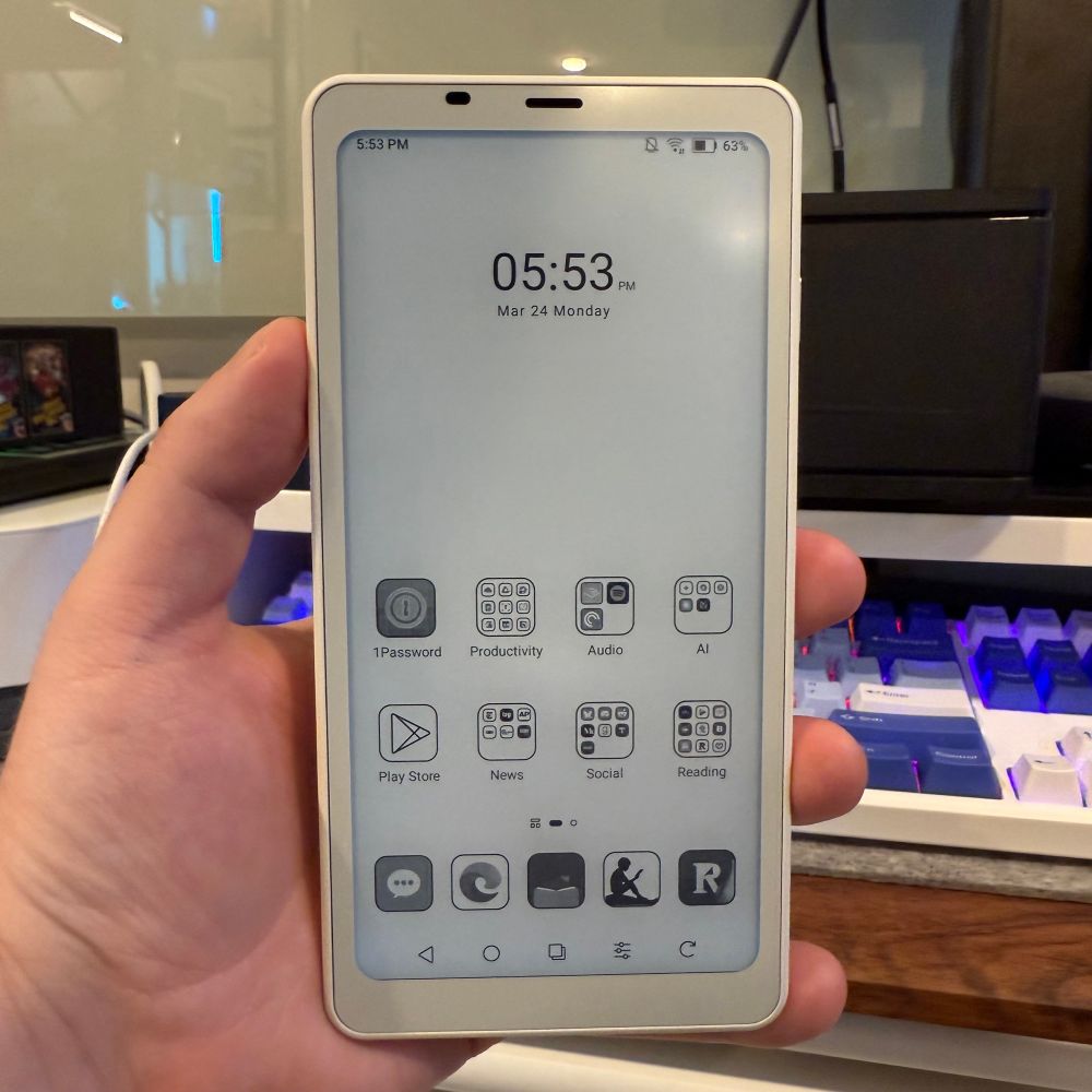 A hand holding a Boox Palma 2 e-ink device displaying a home screen. The time reads ‘05:53 PM’ on ‘March 24, Monday.’ The screen shows app icons organized into labeled folders including ‘1Password,’ ‘Productivity,’ ‘Audio,’ ‘AI,’ ‘Play Store,’ ‘News,’ ‘Social,’ and ‘Reading,’ along with a row of additional apps and navigation icons at the bottom.