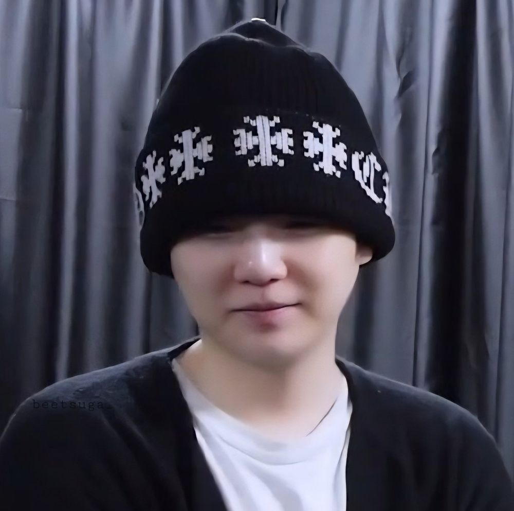 Min Yoongi round and smooth face, smiling with a black beanie covering up to his eyes. He is wearing a white t-shirt and a black cardigan. His face is round and I want to poke his nose 🥹.