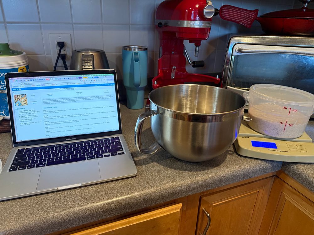 Sourdough starter in a cylinder clear tupperware, large mixing silver mixing bowl and a macbook with a recipe open for Hillary’s Reliable, Good Bread. all on a gray kitchen counter with various appliances behind them. 