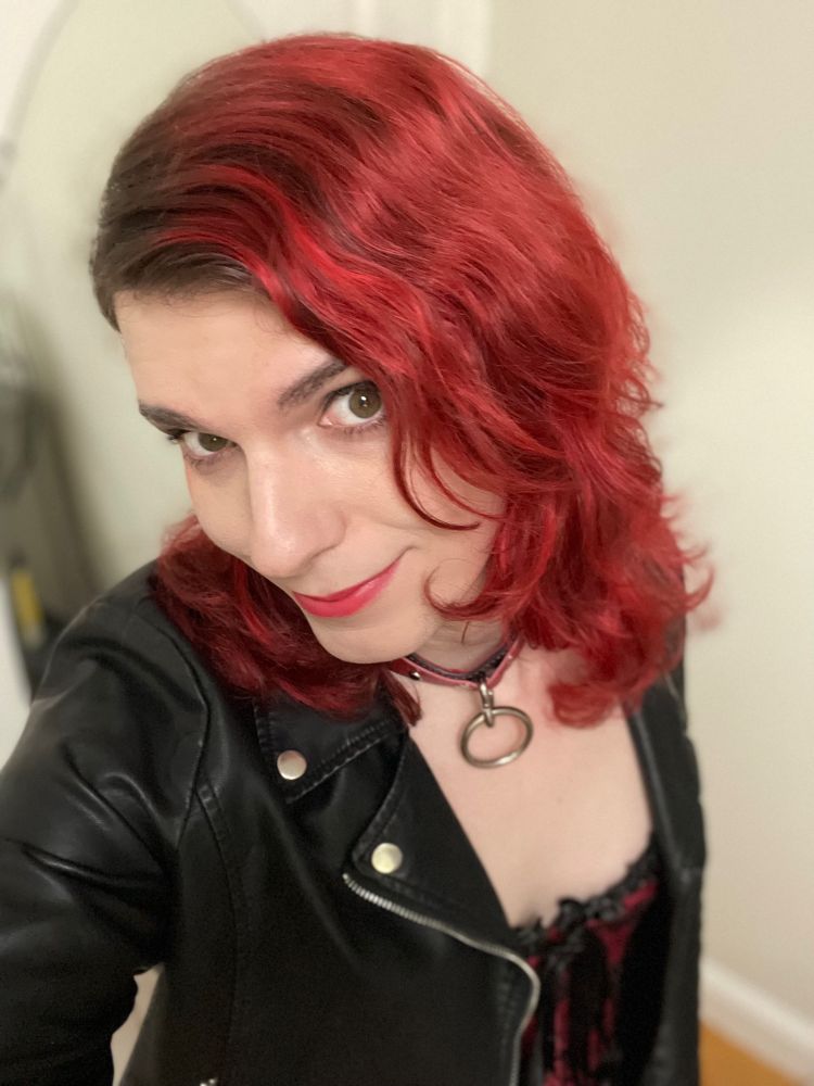 Picture of me, with hair a bright red and roughly at shoulder’s length, a black jacket, black and red corset, black and red leather collar, and a bright red lipstick color 