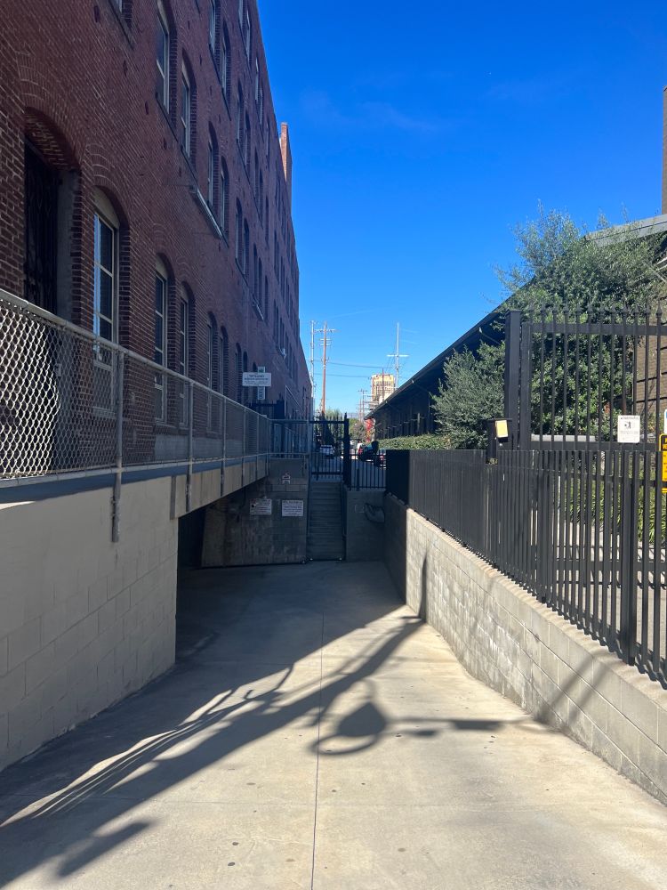 Snapshot of a driveway next to a brick building, which is never to a smaller gated driveway and another building with green trees and shrubbery all around it. The sky is very blue with only a tiny wisp of a cloud.