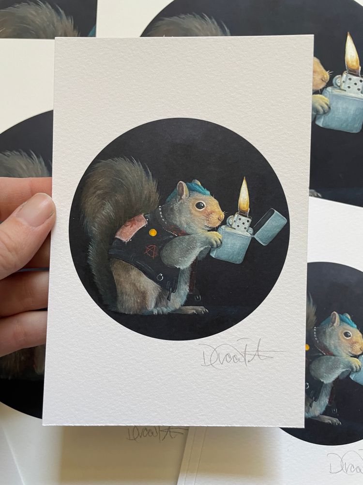 Photo shows a hand holding a print in front of a stack of prints on a table. The print is of an Oil painting of a squirrel in a punk rock leather vest with a green long haircut. The squirrel is holding a lighter and her face is glowing from the light. The rest of the painting is dark and moody.