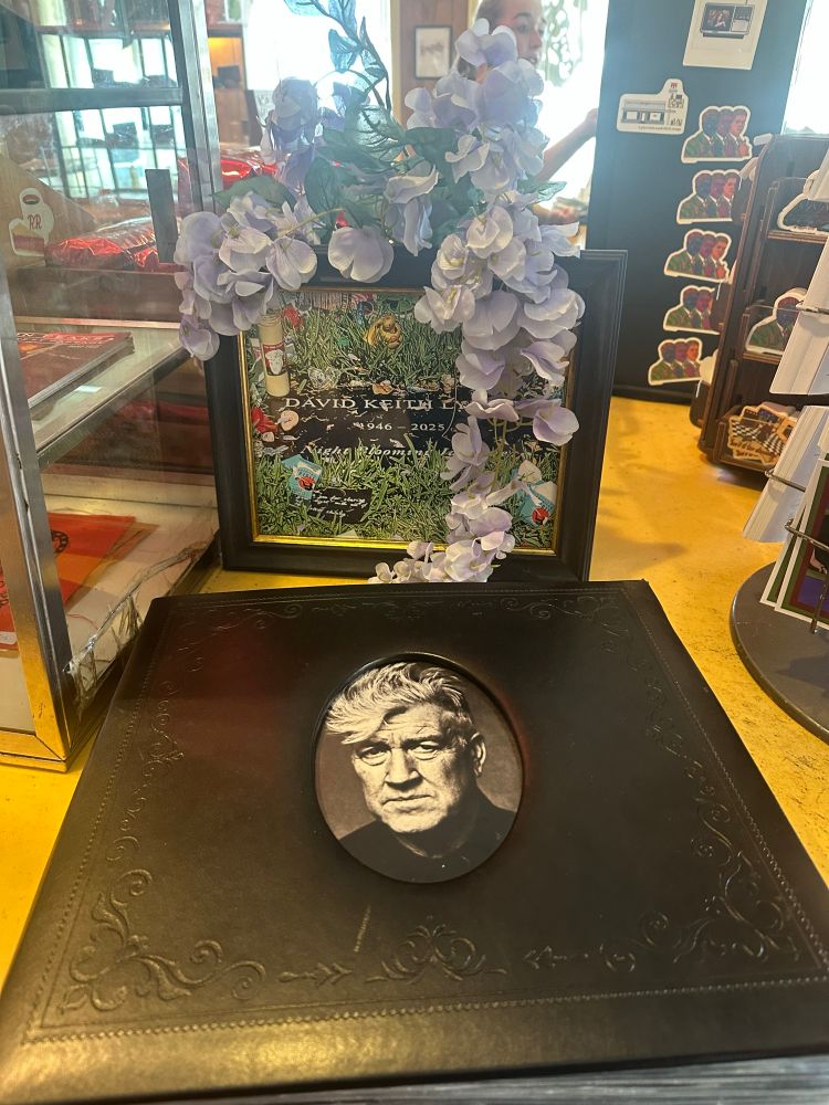 A framed grayscale photo of David Lynch, placed before a photo of his gravestone, which reads “DAVID KEITH LYNCH - 1946–2025 - Night Blooming Jasmine”. The second photo is wreathed in purple flowers. 