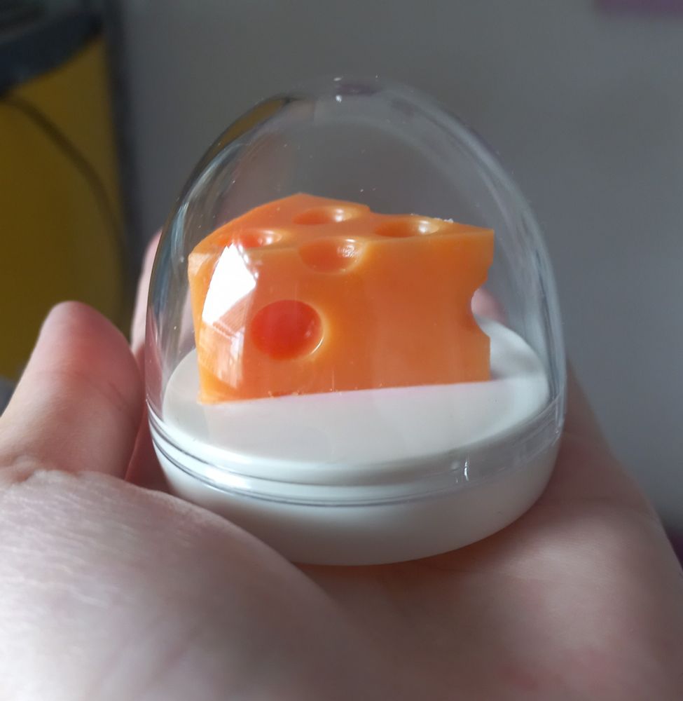 mão com a palma pra cima segurando um lip balm em formato de mini fatia de queijo suíço, que deveria ser amarelo, mas está laranja