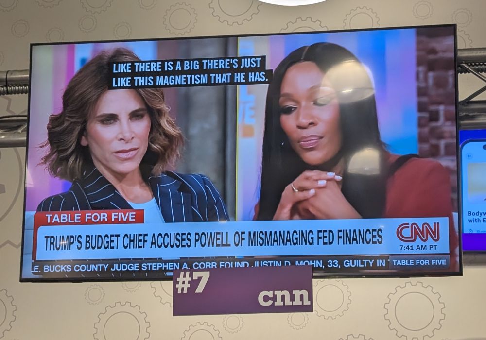 Photo of a Planet Fitness tv airing the CNN program Table for Five with a split screen of "The Biggest Loser" host and noted terrible person Jillian Michaels on the left and CNN Contributor Cari Champion on the right with the chyron reading "Trump's Budget Chief Accuses Powell of Mismanaging Fed Finances"