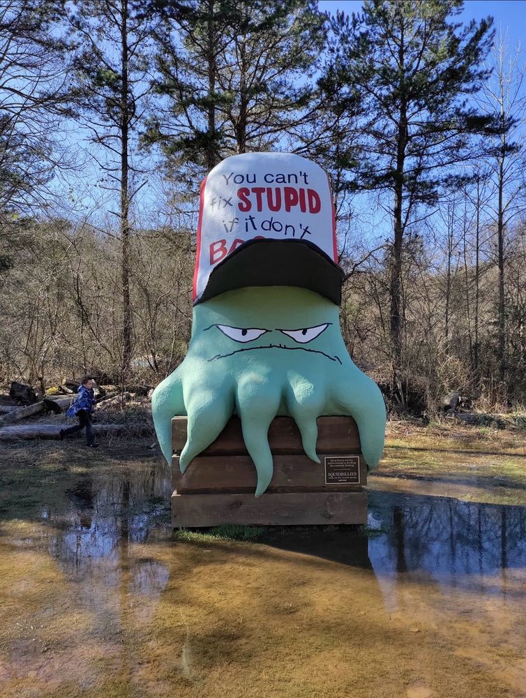 a big statue of early cuyler sitting on an overturned wood crate. his hat says ‘you can’t fix STUPID if it don’t BROKE.’ the surroundings are very forested and marshy