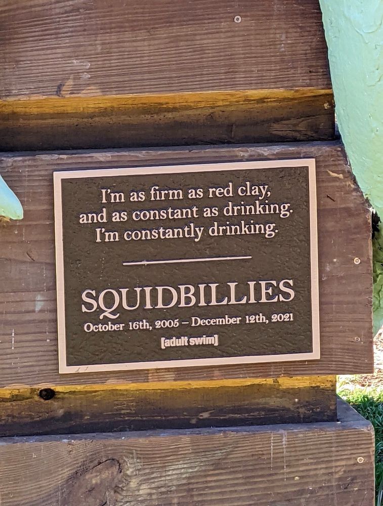 the plaque on the crate. it says 

I'm as firm as red clay, and as constant as drinking.
I'm constantly drinking.
SQUIDBILLIES
October 16th, 2005 - December 12th, 2021
[adult swim]