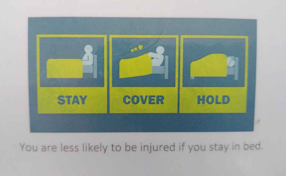 A safety chart graphic from a hotel, instructing that in event of earthquake one should STAY in bed, COVER up with a duvet, and HOLD your head. Text below graphic says "You are less likely to be injured if you stay in bed".