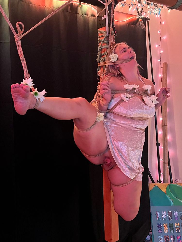 A woman in a shiny dress  is tied upright in bondage against a wooden post. One leg is spread to the side and tied at the ankle exposing her.