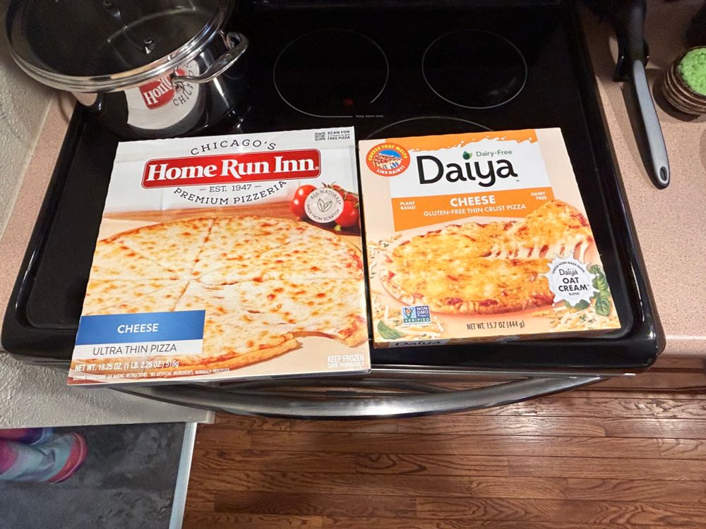 Two frozen pizzas on top of the oven along with a sauce pan. Home Run Inn cheese pizza for me and Daiya dairy-free cheese pizza for my son. 